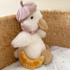 Stuffed toy duck in a basket wearing a pink beret and floral scarf, holding a croissant.
