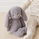 Soft gray plush bunny with long floppy ears and embroidered eyes, sitting beside a child in floral pajamas, lifestyle view.