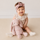Soft pink glitter plush bunny toy by Snuggle Bunnies, held by baby in floral romper and bow headband, front view