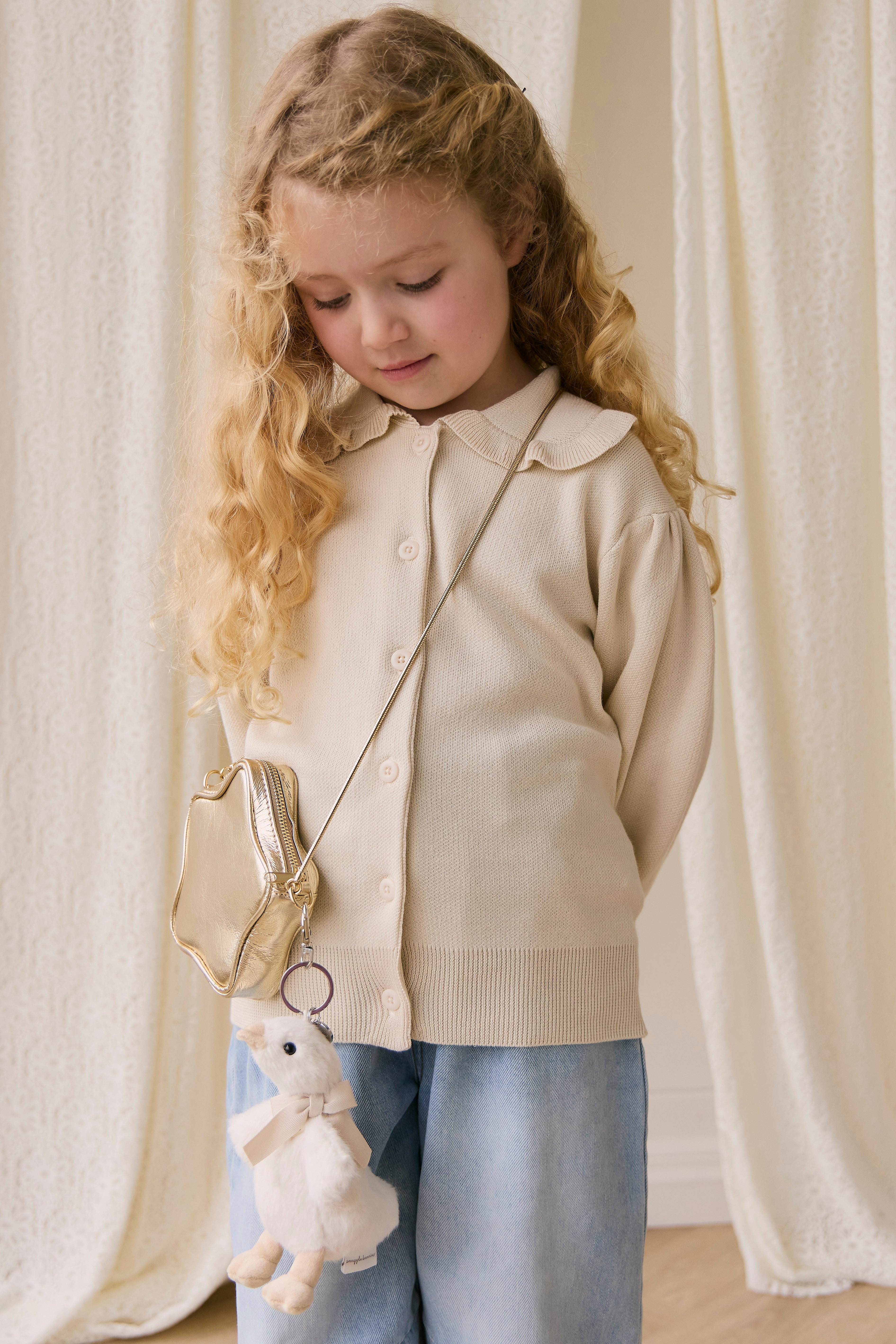 Young girl wearing a beige cardigan holding a teddy bear in a softly lit room.