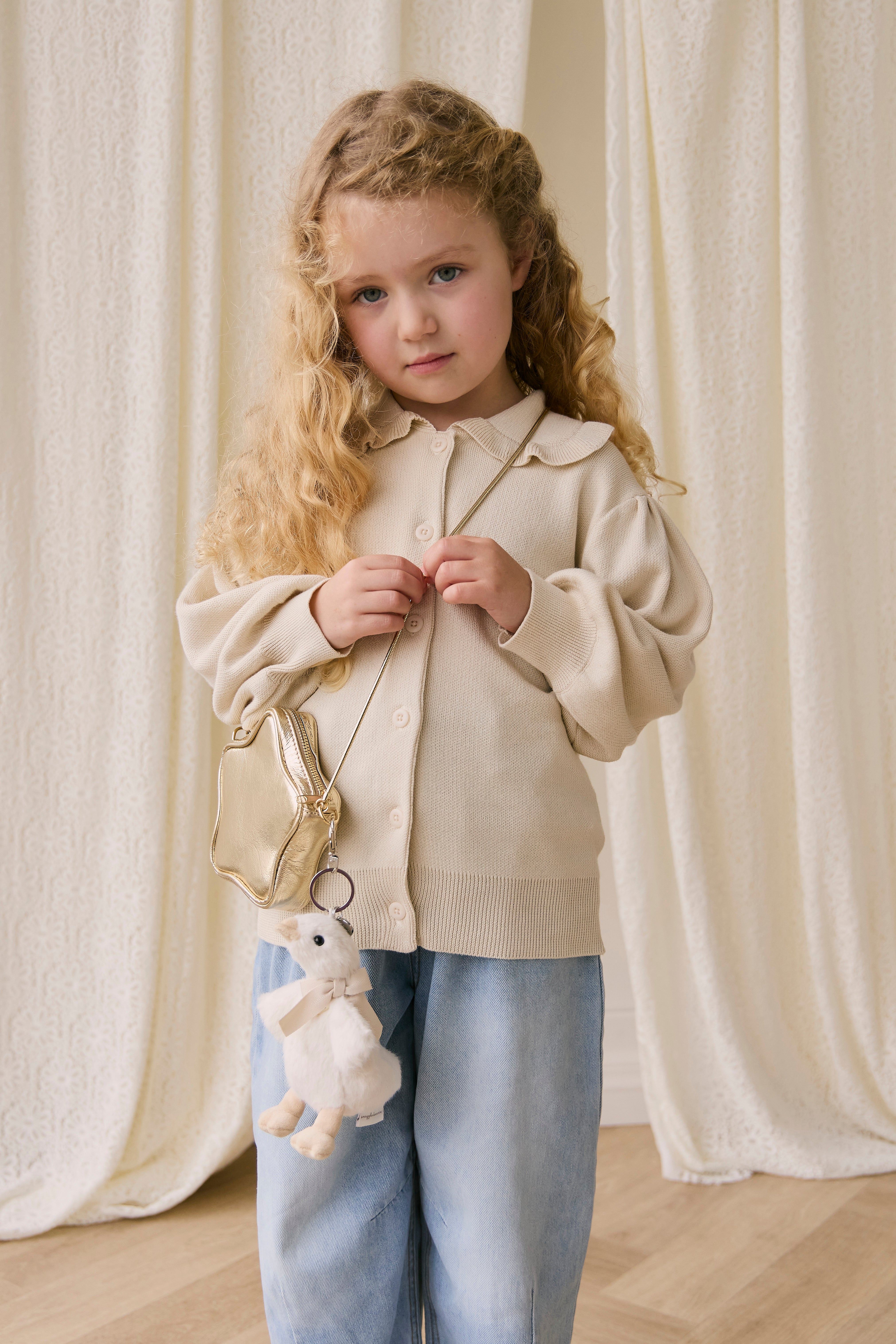 Young girl wearing a beige sweater and light blue jeans, holding a small bag with a plush toy, against a white curtain background.