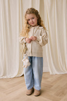 Young girl in a beige jacket and blue jeans holding a white teddy bear against a white curtain background.