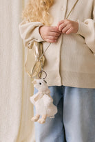 Person holding a plush keychain shaped like a bear with a bow, wearing a beige cardigan and light blue jeans.