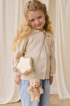 Young girl holding a star-shaped handbag with a teddy bear attached, standing against a white curtain background.