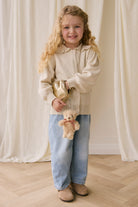 Young girl holding a teddy bear and a gold bag against a white curtain background