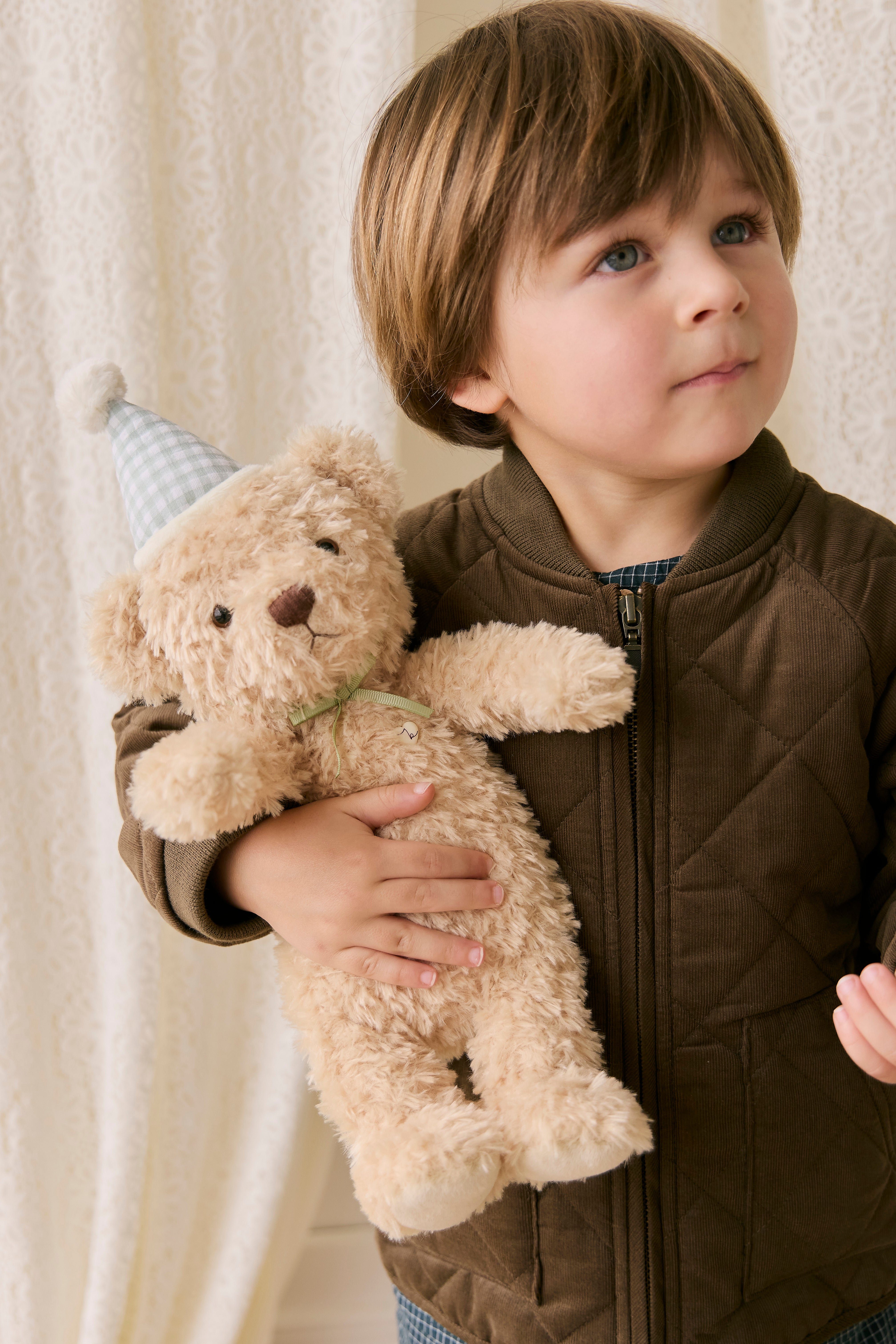 Child holding a teddy bear with a party hat against a neutral background