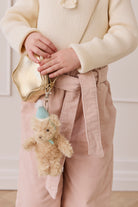 Person holding a gold handbag with a teddy bear charm, wearing a beige coat and pink pants.