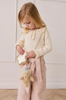 Young girl holding a small handbag with a teddy bear toy, wearing a cream sweater and pink pants.