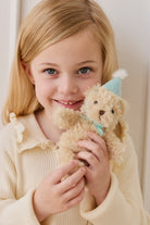 Young girl holding a teddy bear with a party hat against a plain background