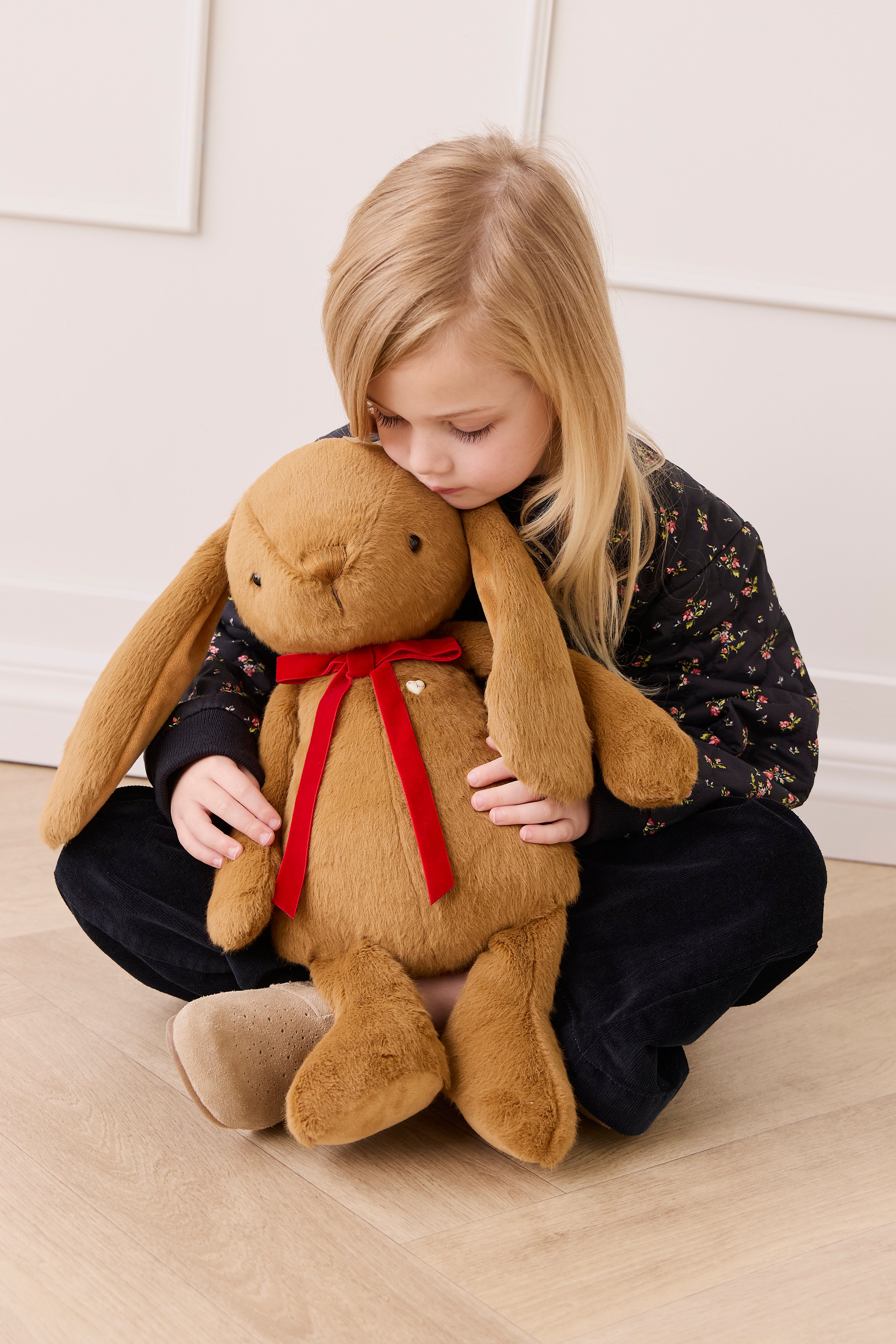 Child holding a large brown plush bunny with a red bow tie on a light wooden floor.