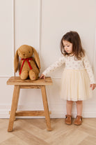 Child in a white dress standing next to a wooden stool with a large plush rabbit toy against a white wall.