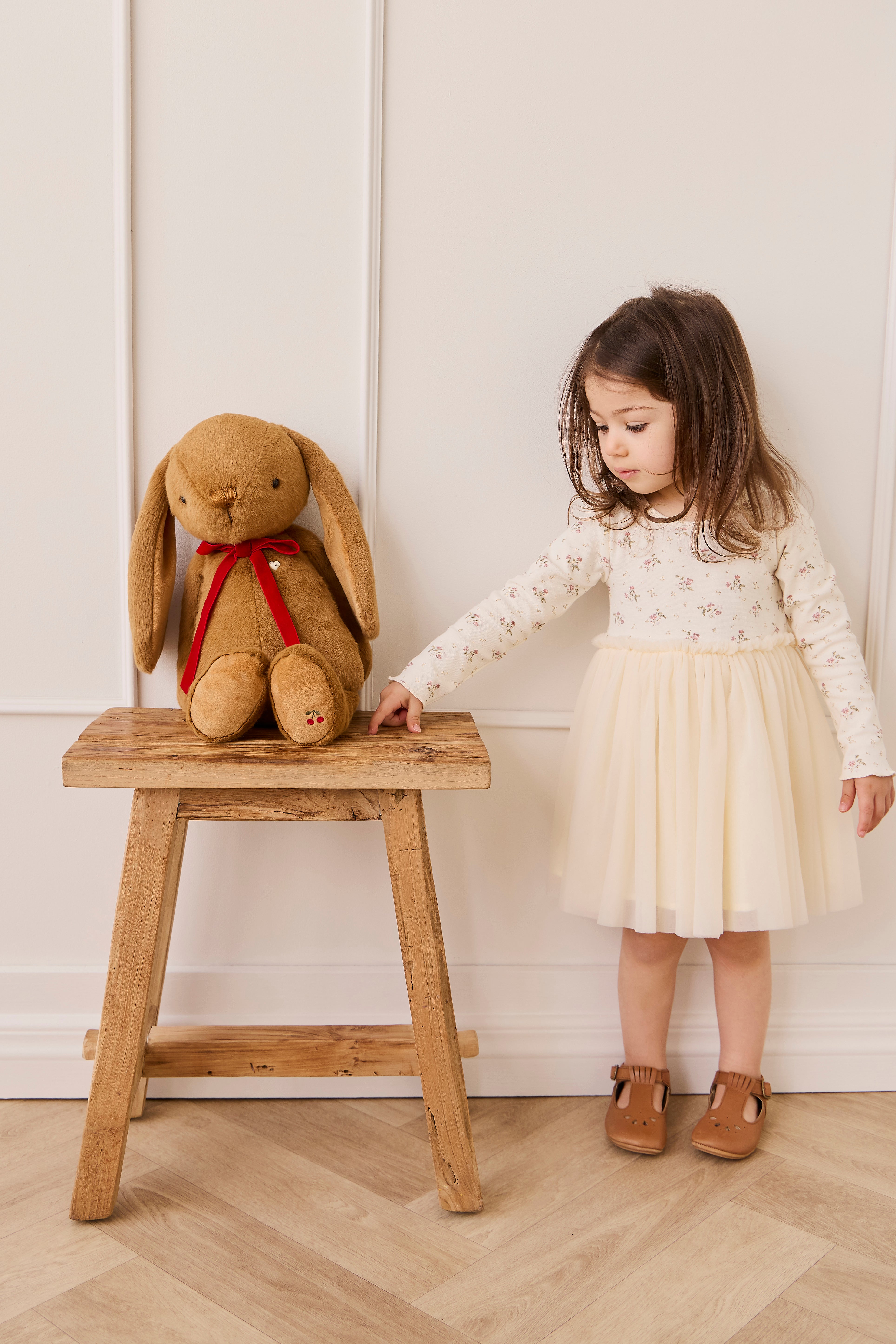 Child in a white dress standing next to a wooden stool with a large plush rabbit toy against a white wall.
