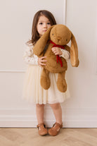 Young girl holding a large brown teddy bear with a red bow against a white wall.