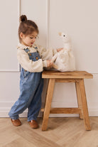 Child in denim overalls interacting with a plush toy on a wooden stool against a white wall.