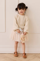 Young girl in a cream sweater and skirt holding a teddy bear against a white wall.