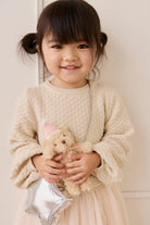 Young girl holding a teddy bear wearing a beige sweater and skirt against a white background