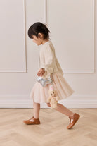 Young girl in a beige outfit with a silver clutch and teddy bear toy, standing on a wooden floor.