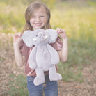 A smiling girl holds a gray plush elephant, wearing a blush-colored velvet shirt and blue jeans outdoors.