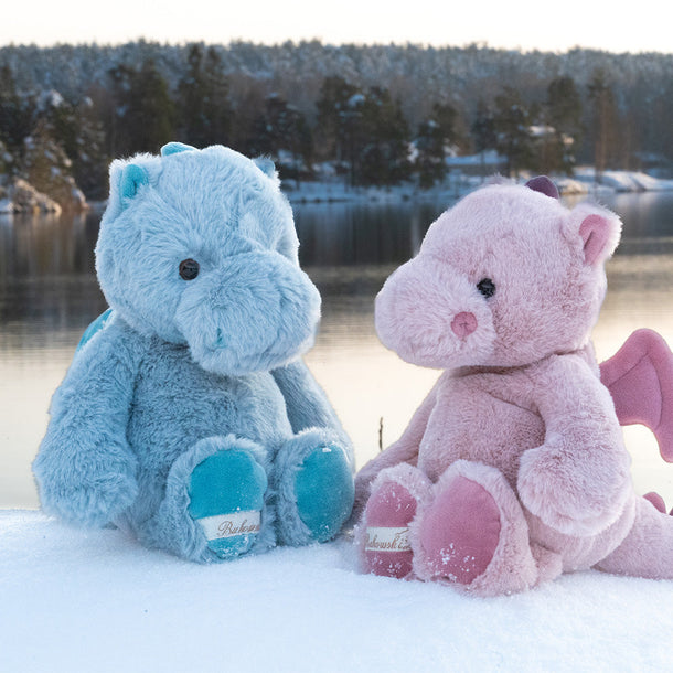 Two teddy bears, one blue and one pink, sitting on a snowy surface with a lake and trees in the background.