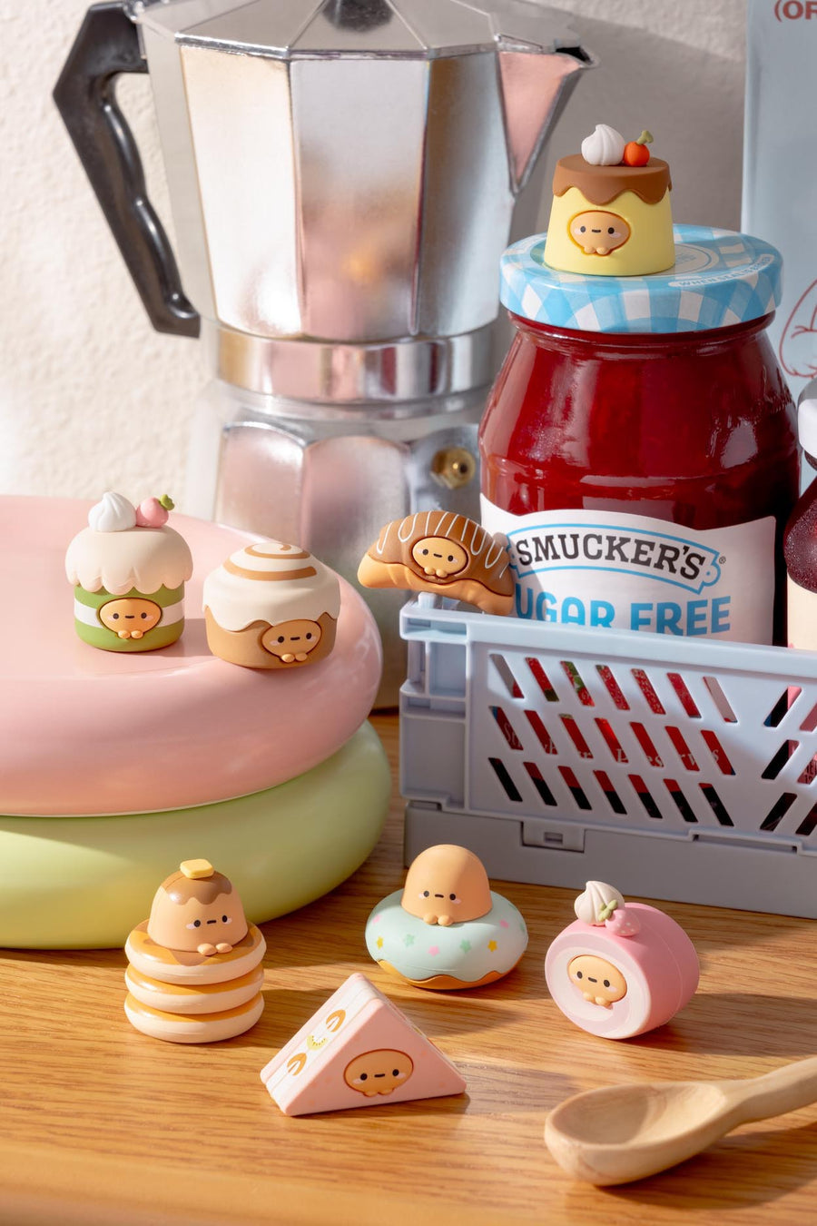 Small figures of pastries and a jar of jelly on a wooden surface with a coffee maker in the background.
