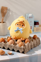 Plush toy resembling a yellow chick with a blue bow tie sitting on a cardboard egg carton filled with eggs.