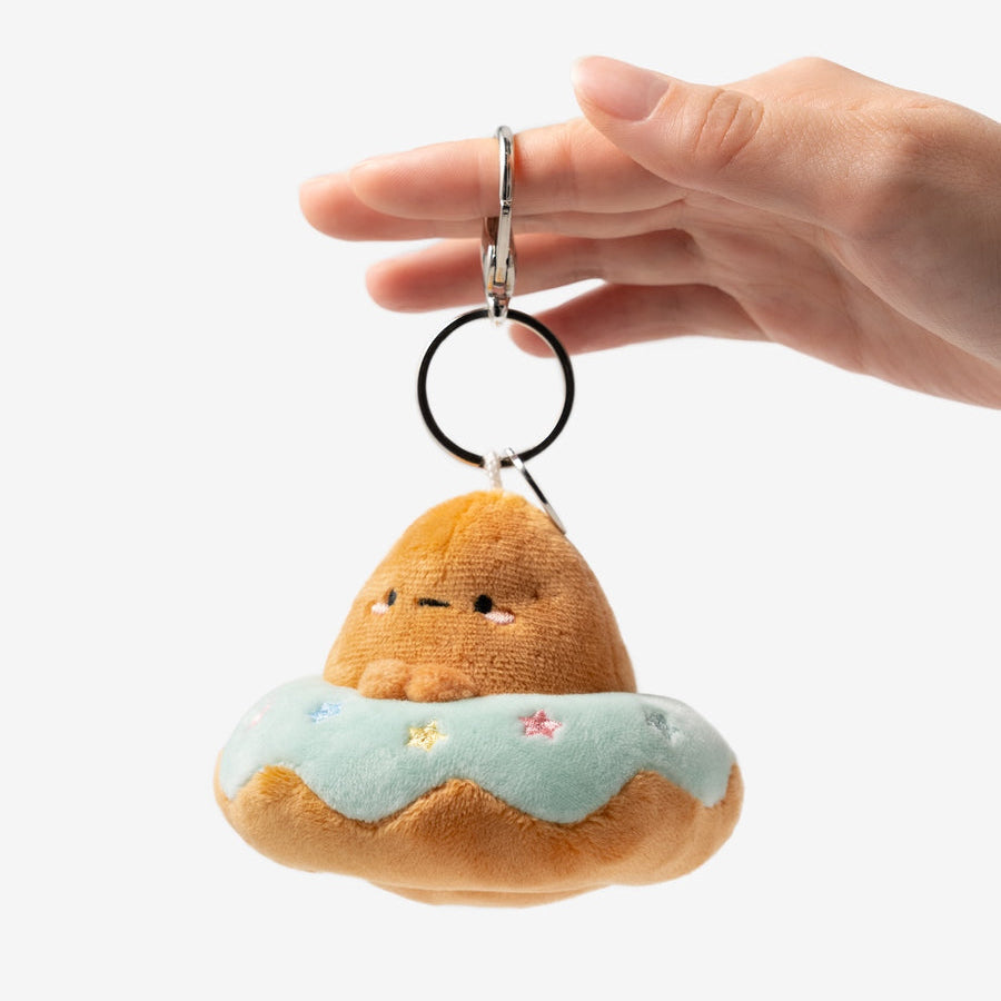 A hand holding a small plush keychain shaped like a cute brown character sitting on a light blue donut with tiny colorful flower details, against a plain white background.