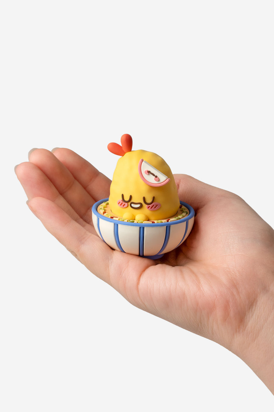 Hand holding a small figurine of a yellow creature with a red comb and a blue bowl.