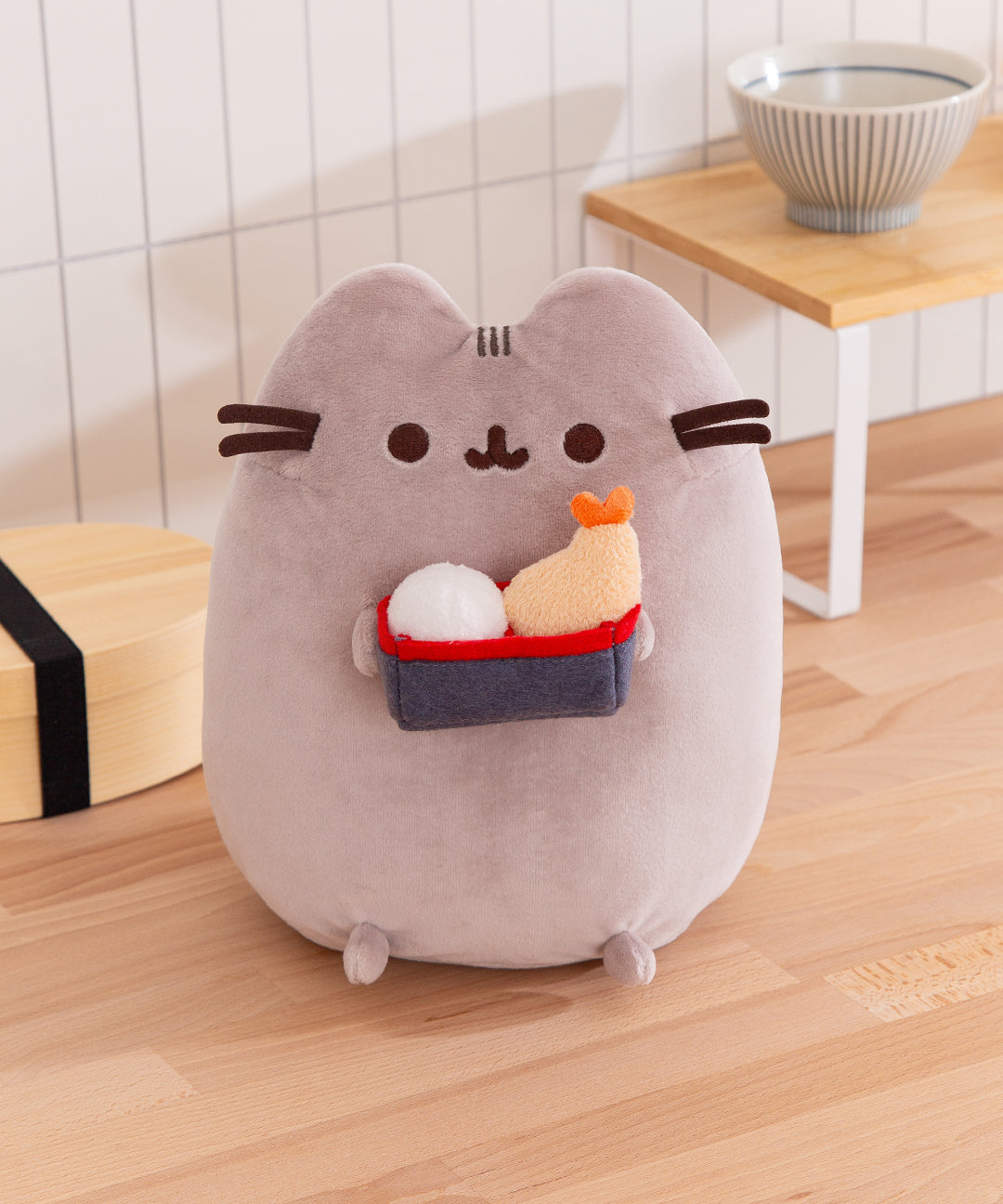 Plush toy shaped like a cat holding sushi on a wooden floor.