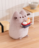 Plush toy resembling a cat holding a small box with a fish on a wooden floor.