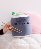 Plush toy shaped like a piece of sushi with a face, held by a hand with chopsticks on a soft surface.