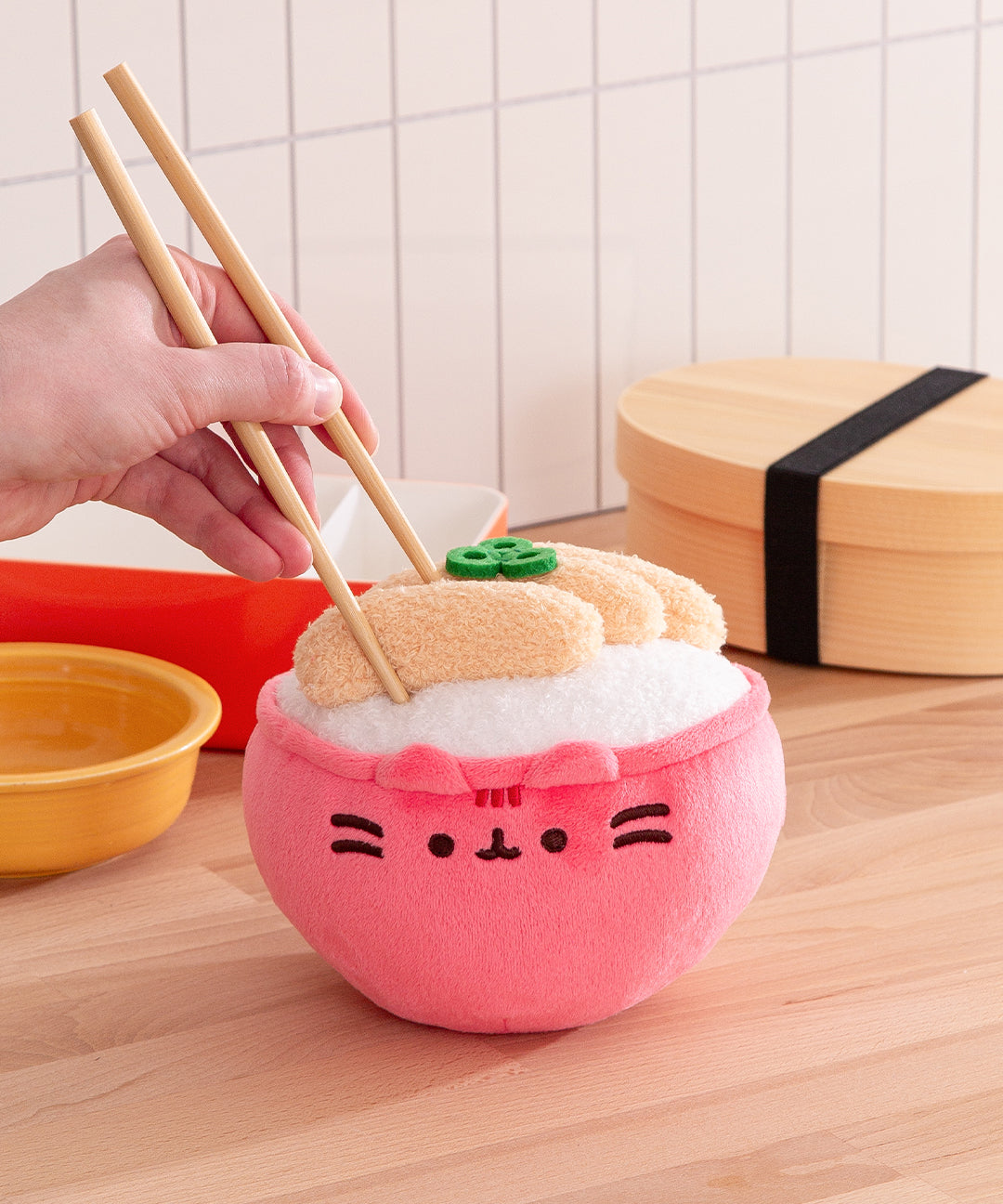 Pink plush toy resembling a bowl of sushi with a hand holding chopsticks over it, on a wooden surface.