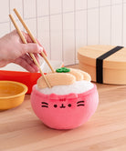 Pink plush toy resembling a bowl of sushi with a hand holding chopsticks over it, on a wooden surface.