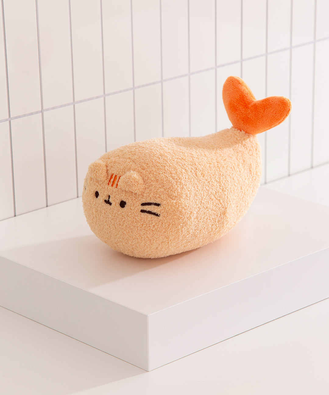 Plush toy resembling a cat with an orange tail on a white surface with a tiled wall background