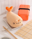 Plush toy shaped like a fish on a plate with an orange bento box and bamboo mat in the background.