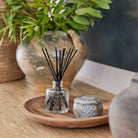 A small glass diffuser with black sticks on a wooden tray, surrounded by decorative items.