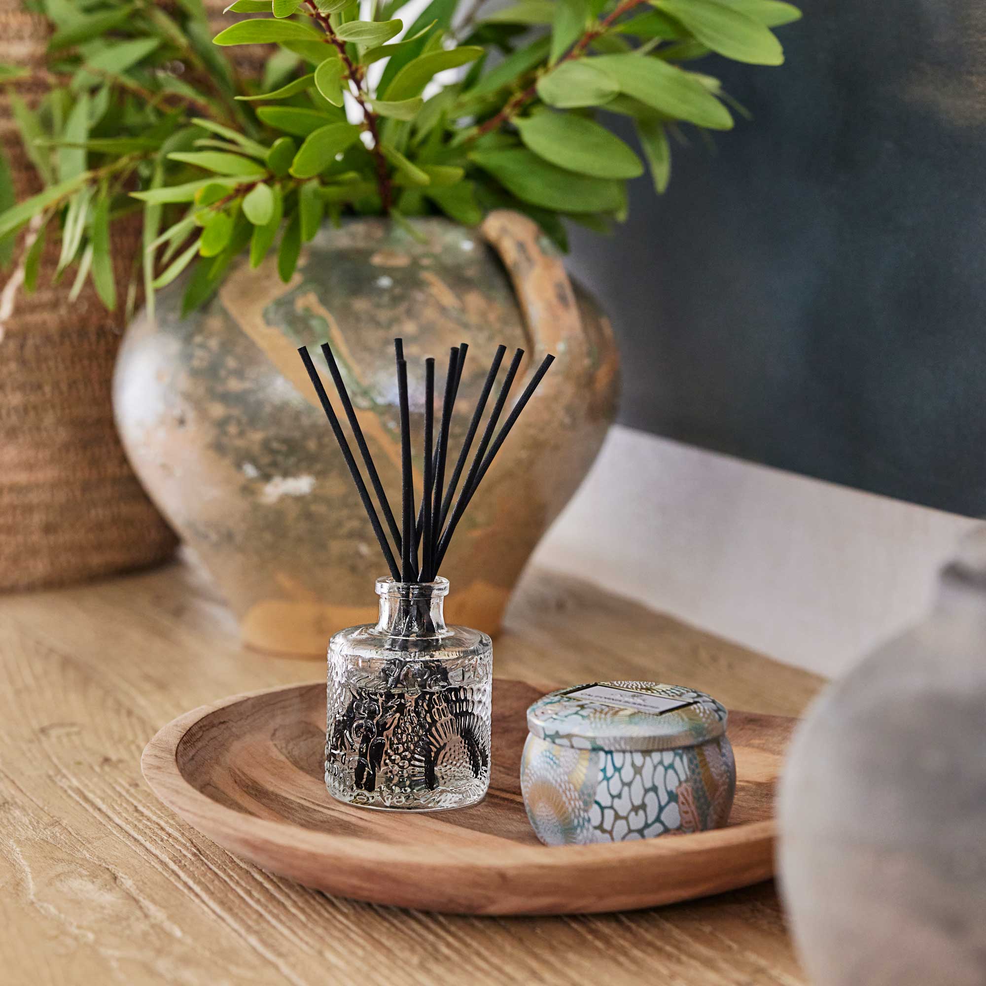 A small glass diffuser with black sticks on a wooden tray, surrounded by decorative items.