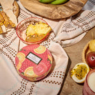 Decorative pink tin with fruit design on a patterned cloth with fruits and a bowl in the background.