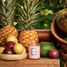 Pink candle with a label surrounded by pineapples, apples, and avocados on a wooden surface.