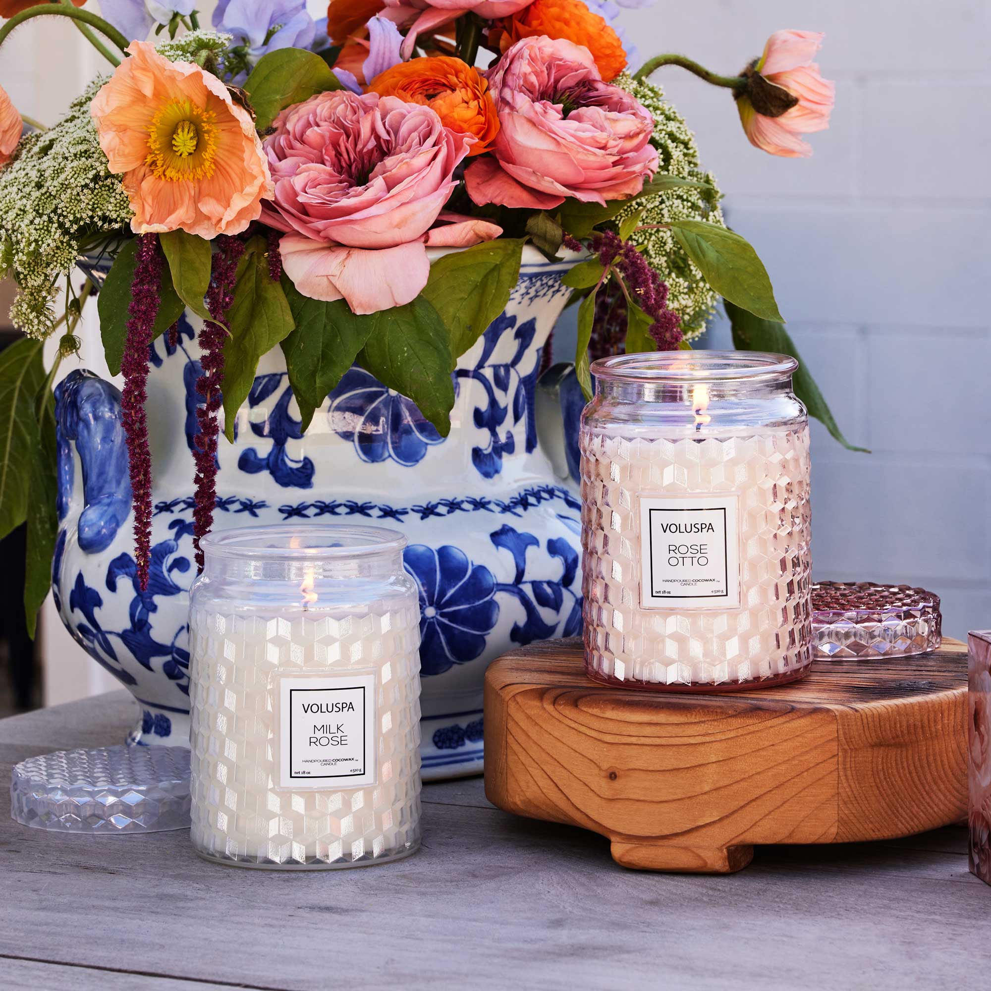 Two Voluspa candles on a wooden coaster with a floral arrangement in the background.
