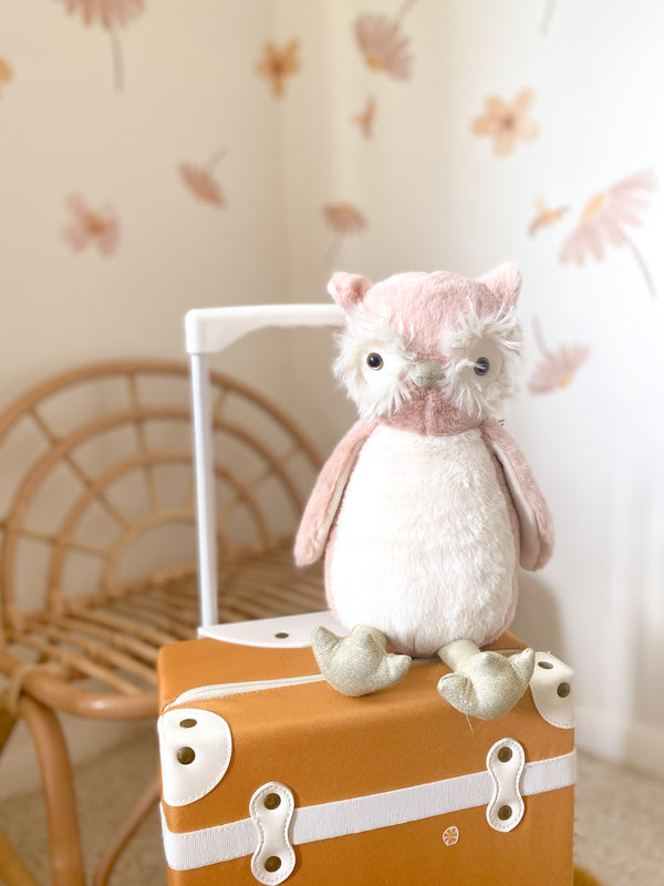 Stuffed owl toy sitting on a brown suitcase with white details, next to a wicker chair.
