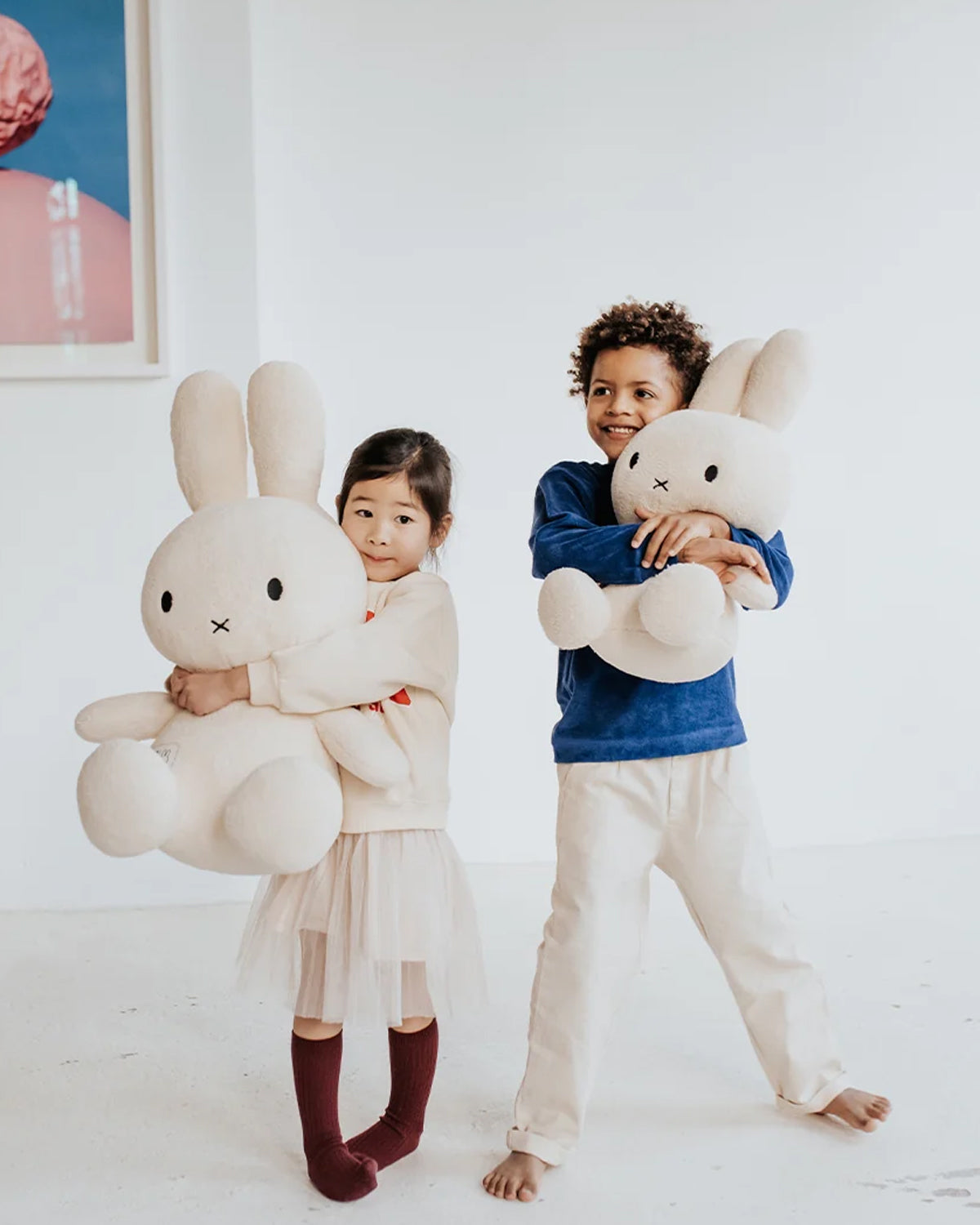 Two children holding large plush bunnies in a room with a white wall and framed picture.