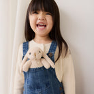 Child wearing denim overalls holding a plush bunny toy against a plain background