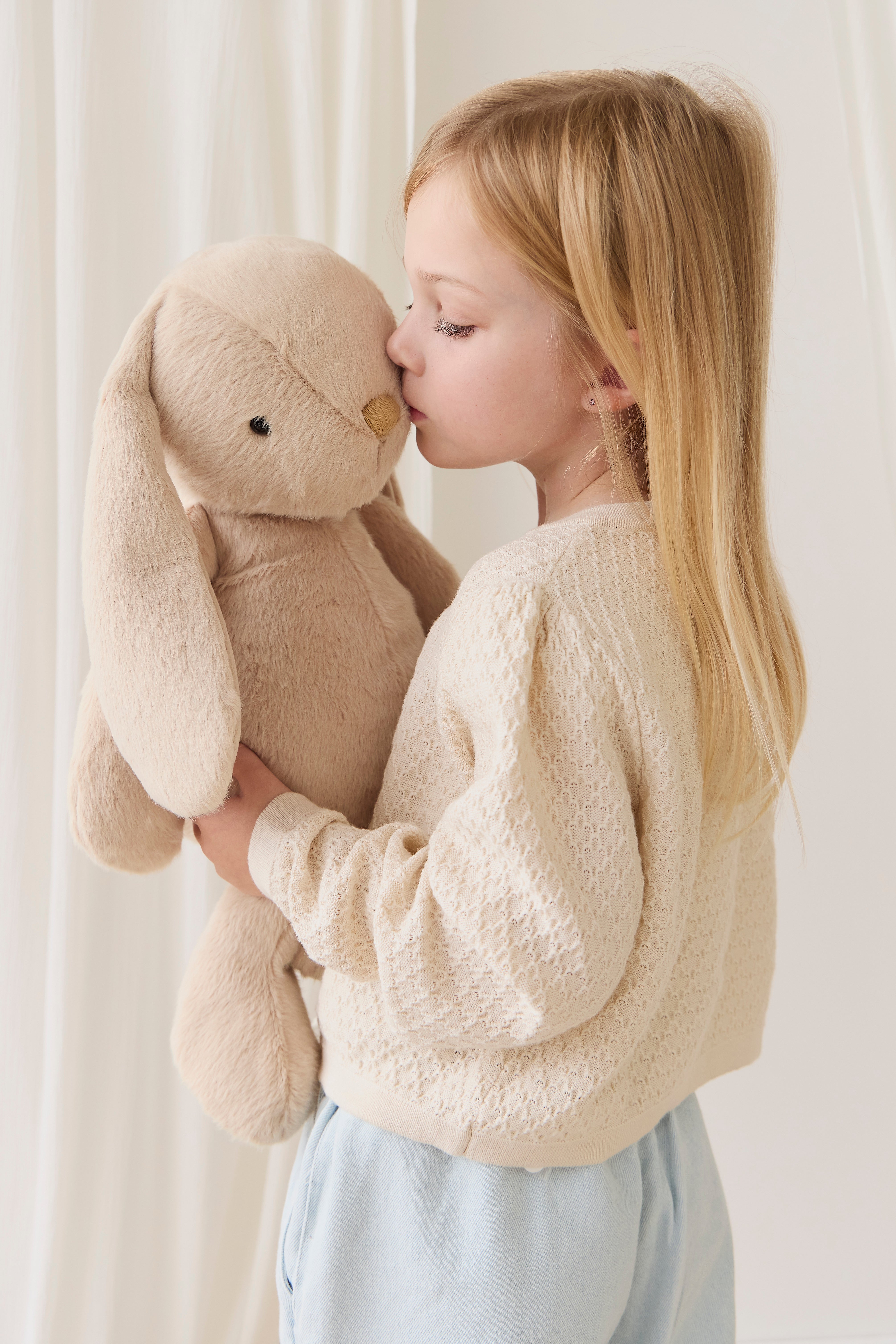 Young girl holding a beige stuffed bunny against a white curtain background