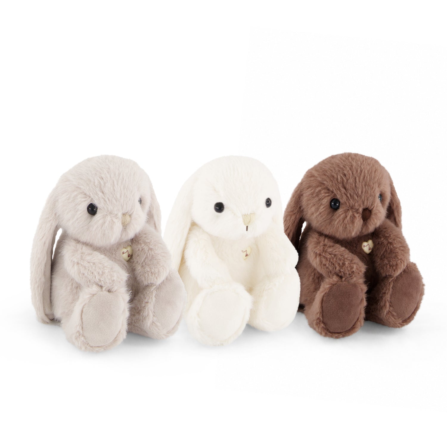 Three plush bunny toys arranged side by side on a white background: one light beige, one white, and one medium brown, each with long floppy ears, black eyes, a small gold bell, and a tiny beige stitched nose.