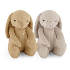 Two plush bunny toys side by side on a white background, one in tan and one in light gray, each with long floppy ears, black bead eyes, a stitched smile, and a small gold heart on the chest.