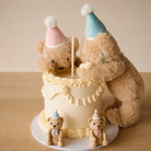 Soft tan plush teddy bear wearing blue party hat with white pom-pom, positioned beside cream frosted birthday cake with ruffled icing, accompanied by smaller matching bears in pink and blue hats, front view.
