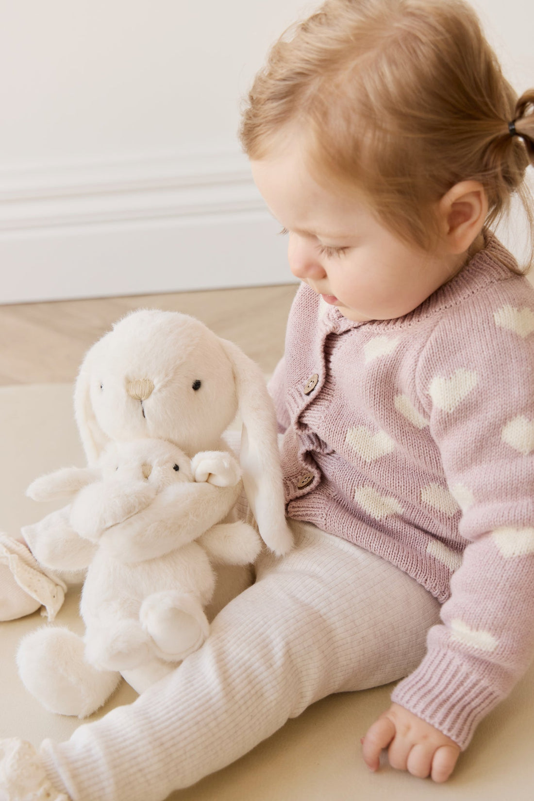 Child in a pink sweater with a plush toy on a light wooden floor.