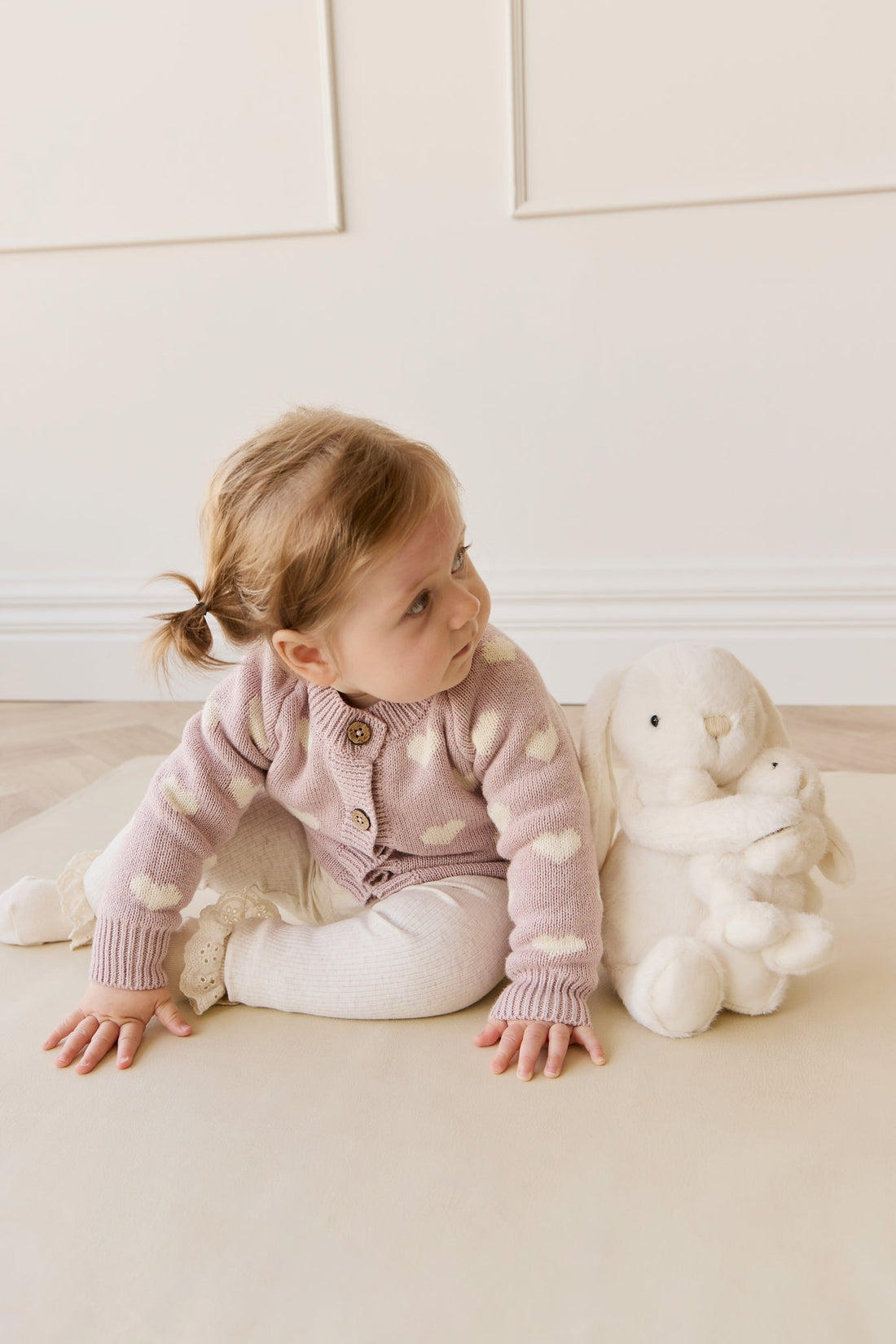 Child in a polka dot cardigan sitting on a light-colored floor with a white teddy bear.