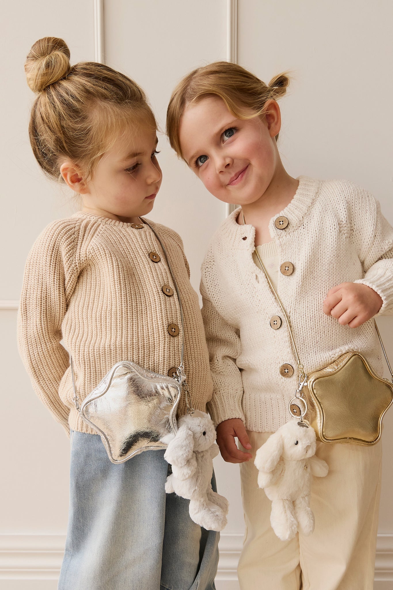 Two young girls pose together in a studio setting, wearing knitted cardigans with wooden buttons and metallic star-shaped bags with stuffed bunny charms. One girl has a bun and is wearing denim pants and a silver bag, while the other girl has a half updo and is wearing cream-colored pants and a gold bag.