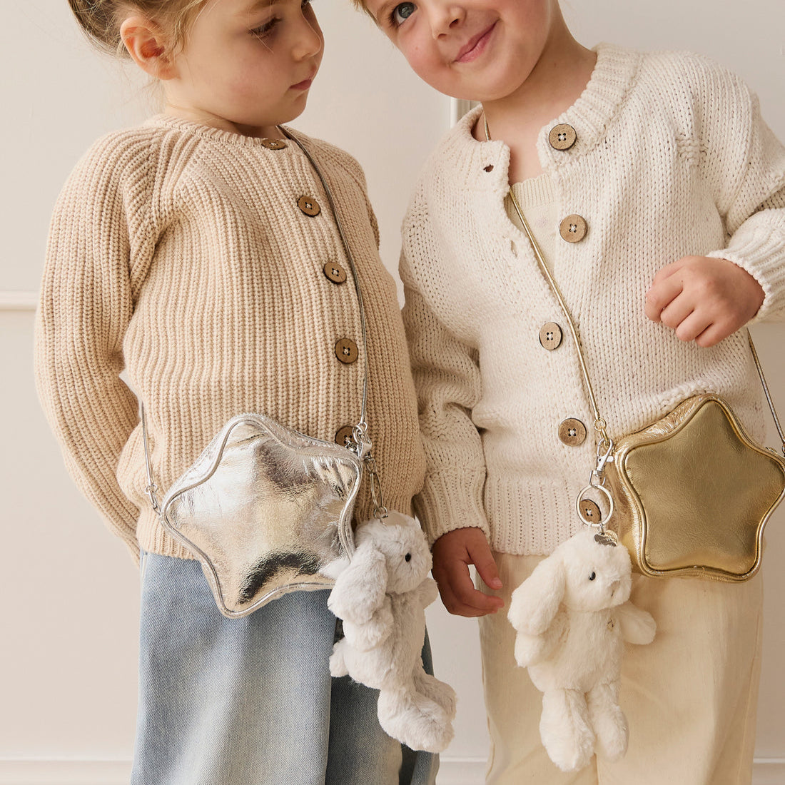 Two young children wearing cardigans and carrying star-shaped purses with plush rabbit keychains.